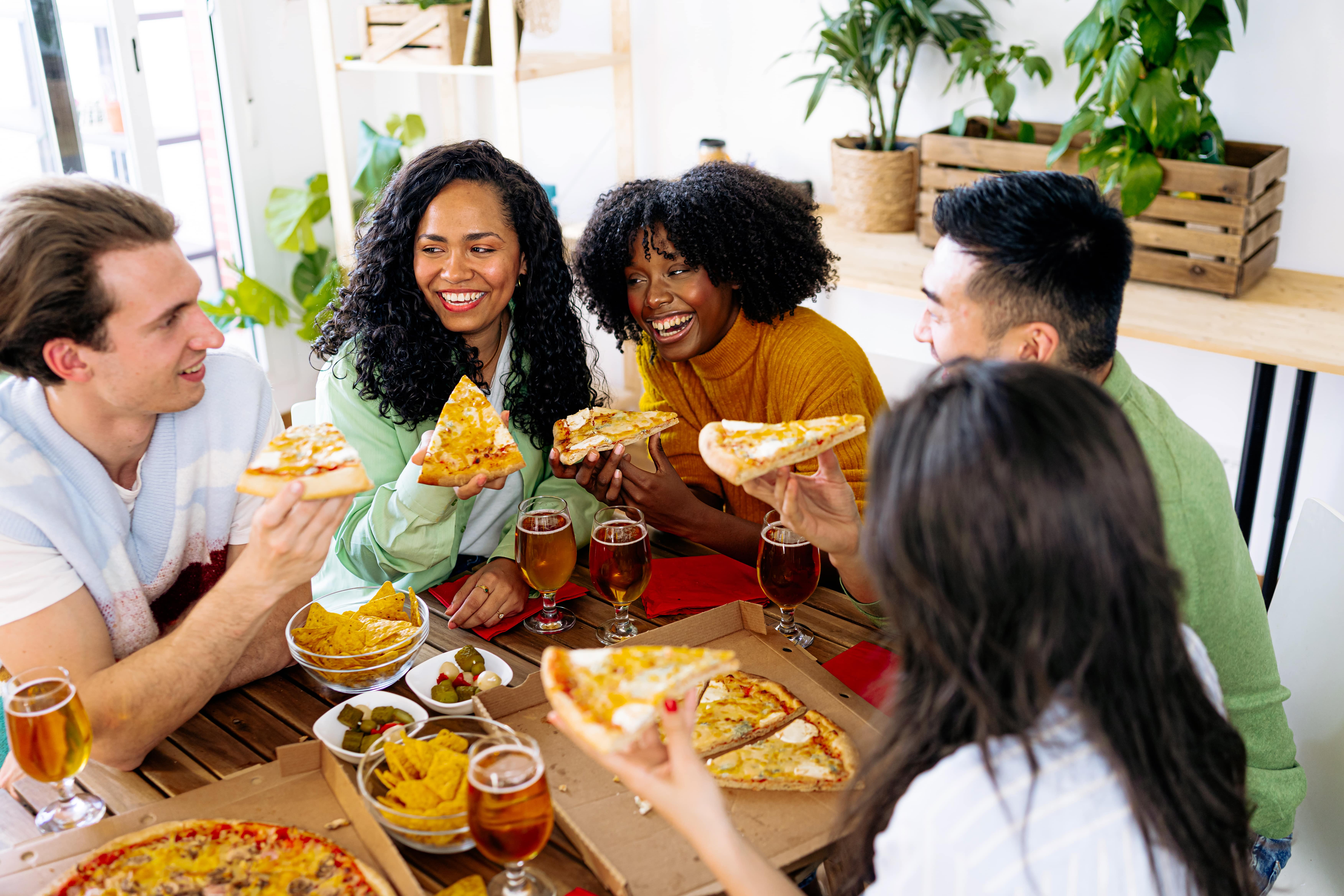 Friends gathered around a table enjoying slices of pizza, with glasses of beer, bowls of chips, and small side dishes in a cozy setting. The group is smiling and laughing, creating a lively and fun atmosphere perfect for a game night or casual gathering.