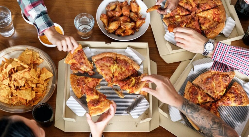 A group of friends gathered around a table, enjoying a lively pizza party. The table is spread with delicious food, including pizza slices, nachos, chicken wings, drinks, and dipping sauces.