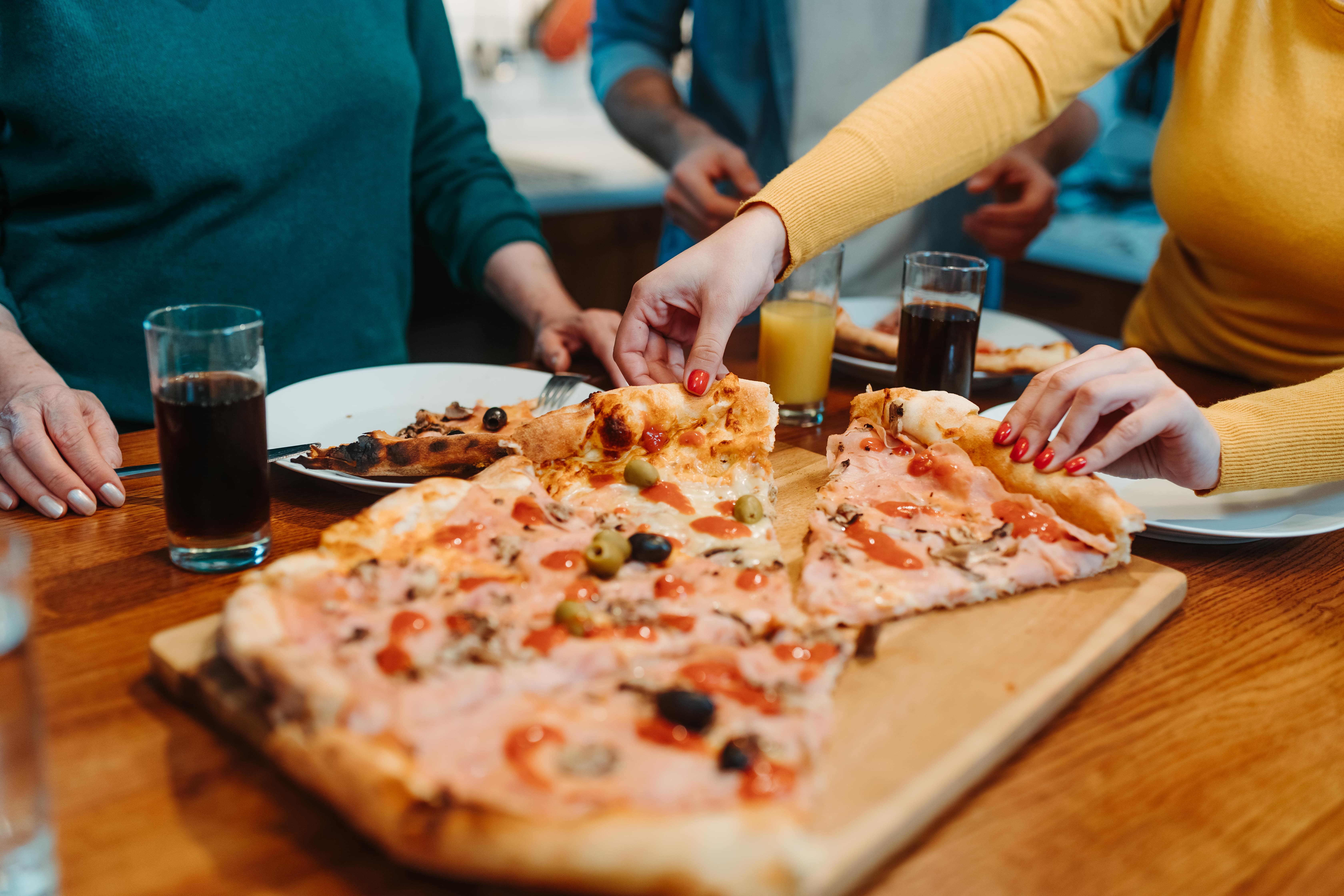 A group of people gathered around a table, sharing a large pizza. There are drinks and plates on the table, suggesting a casual and enjoyable meal.