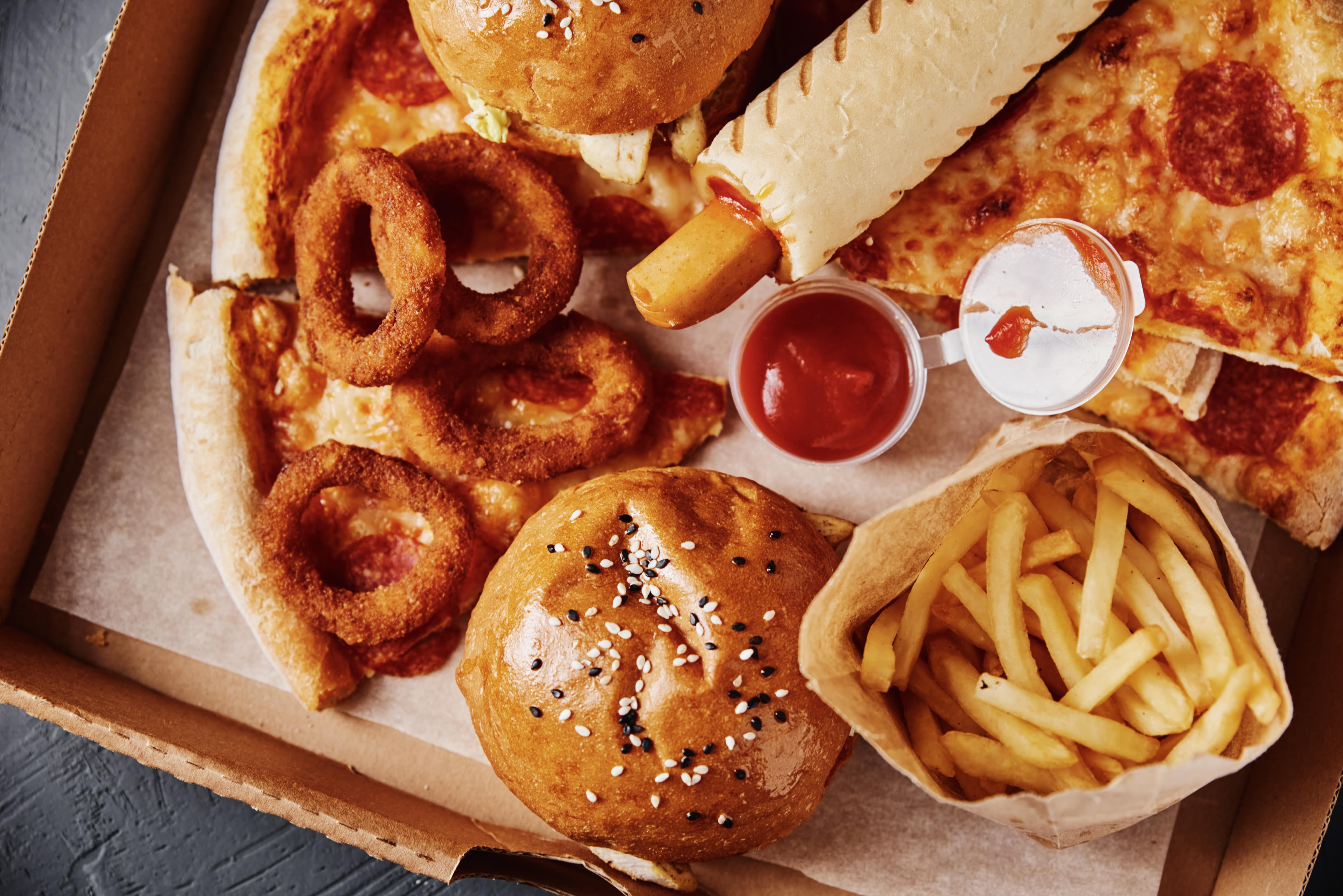 A variety of fast food items, including pizza, burgers, onion rings, and fries, arranged in a takeout container.