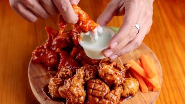 Hand dipping crispy chicken wings into ranch sauce with carrot sticks.